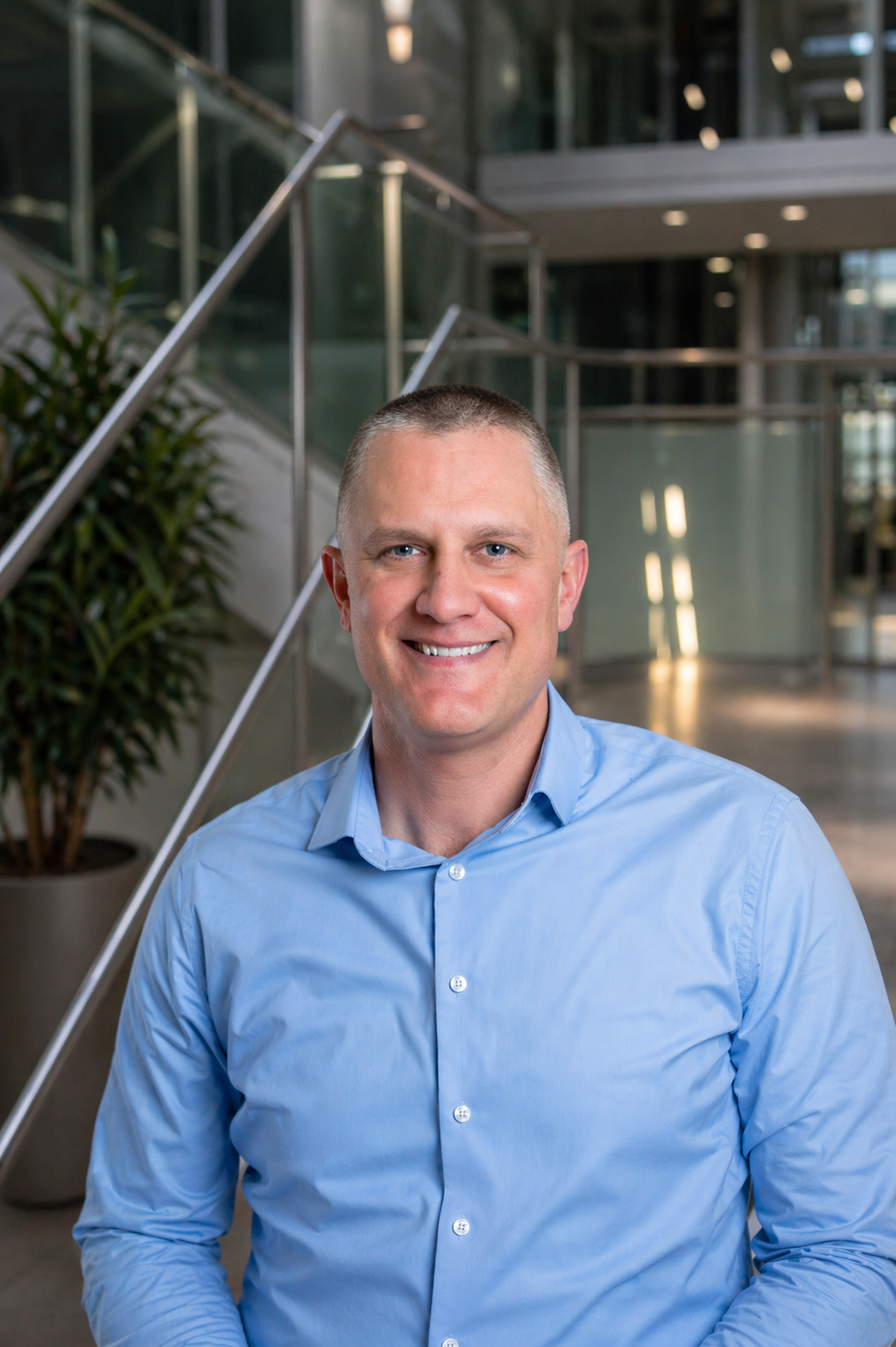 Smiling man in a blue shirt stands in a modern office lobby.