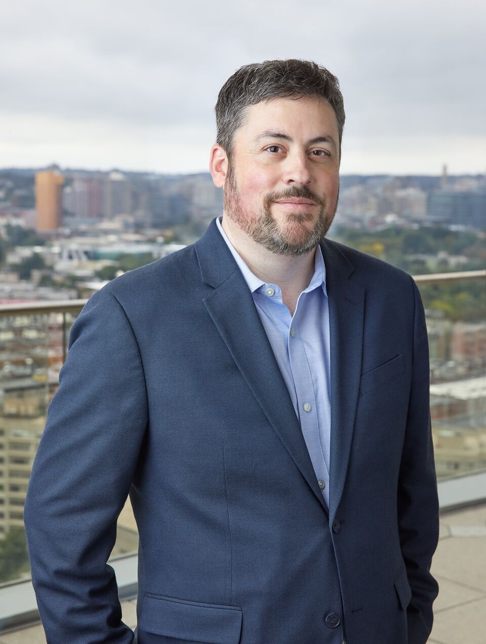 Man in a suit standing outdoors with a cityscape in the background.