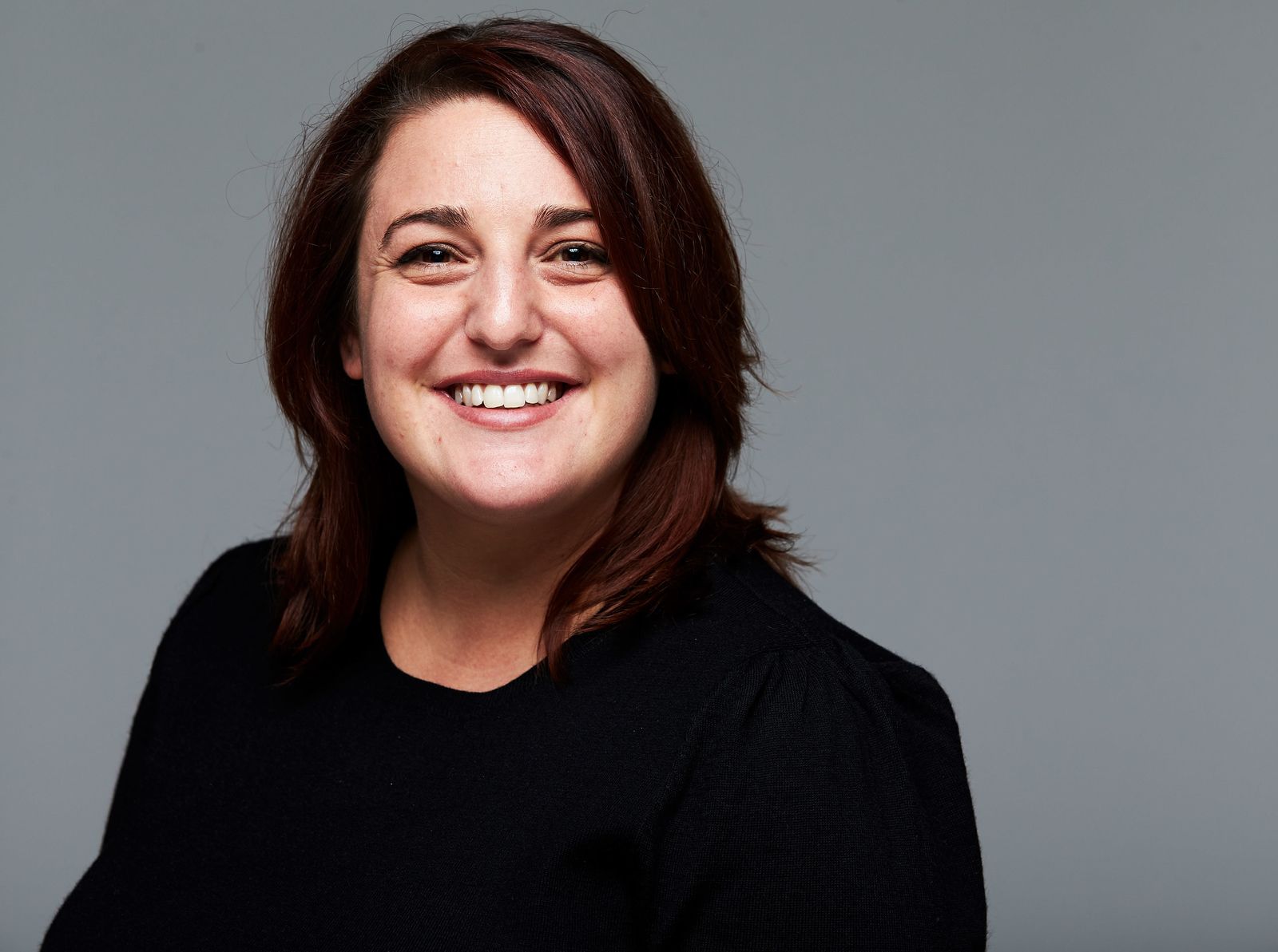 Smiling woman with brown hair wearing a black shirt against a gray background.