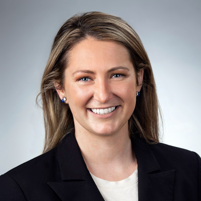 A smiling woman in a black blazer with light hair and earrings.