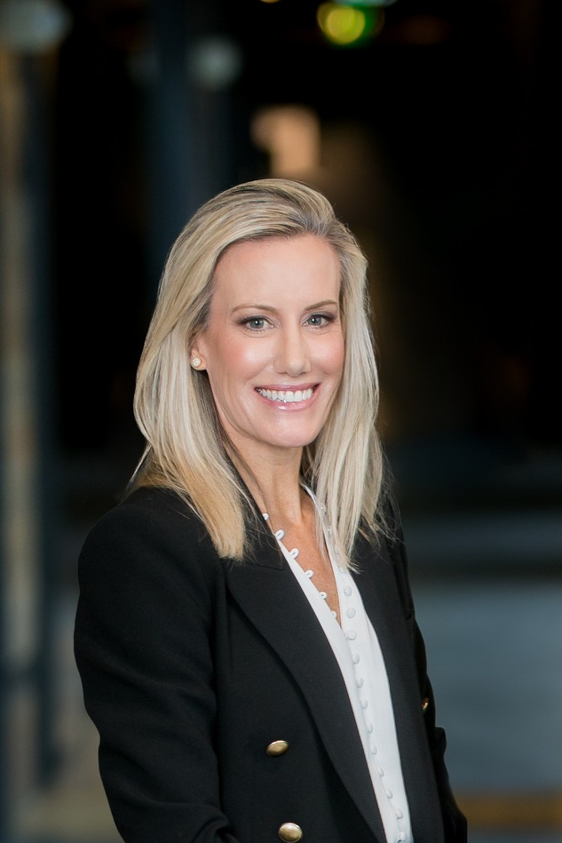 Portrait of Katie Santarelli smiling, wearing a black blazer in a softly lit setting.