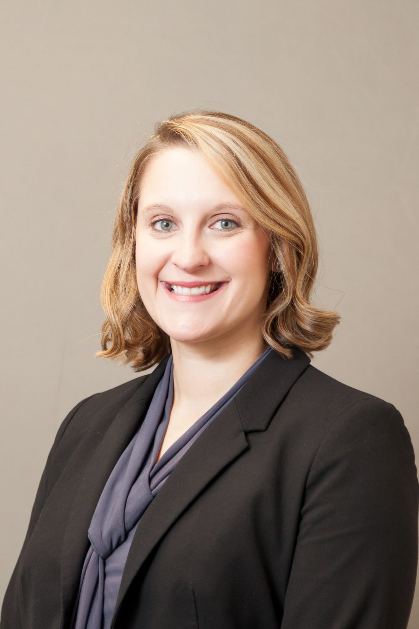 Professional portrait of a woman smiling, wearing a blazer and scarf, with a neutral background.