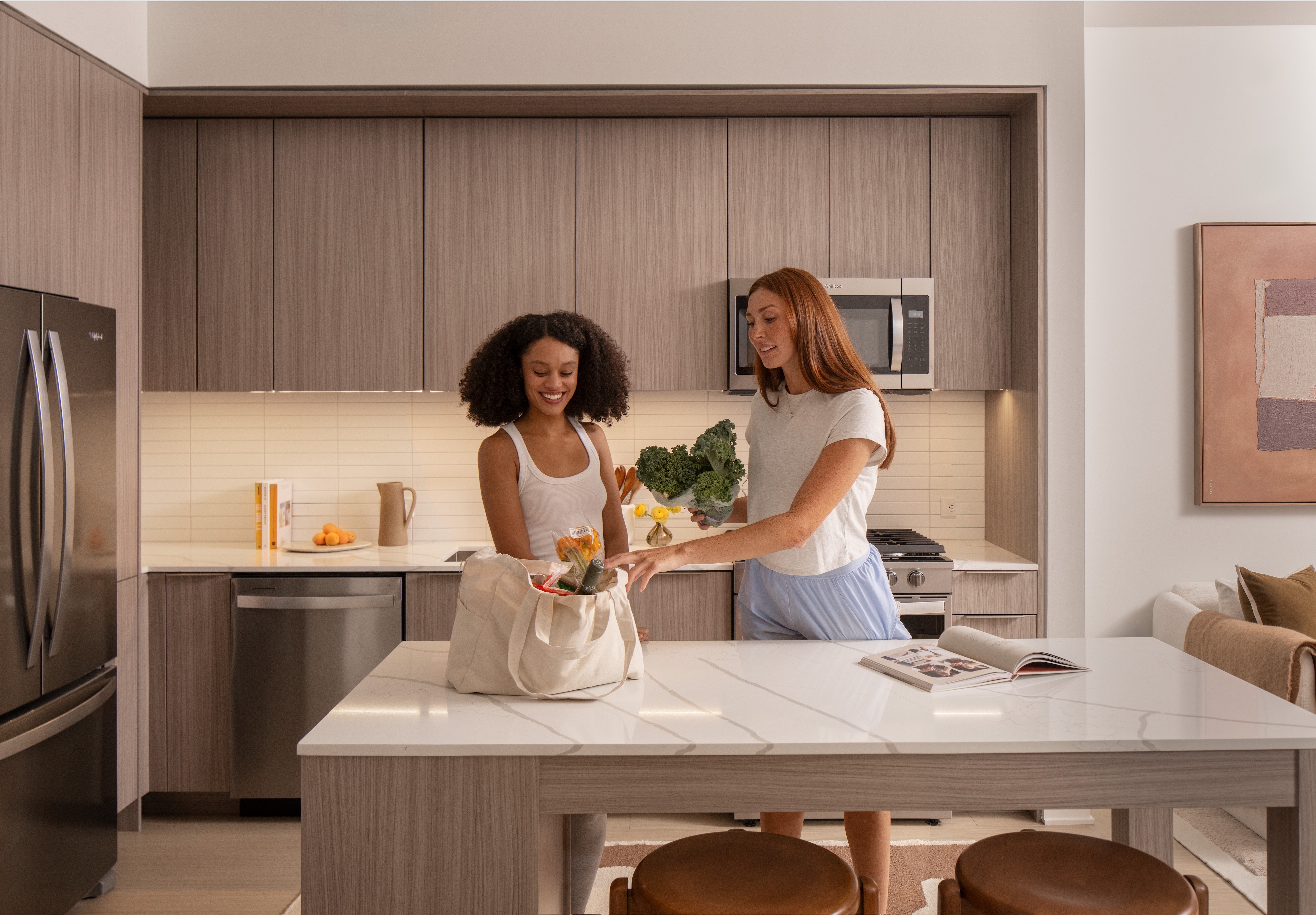 Two women interact in a modern kitchen with light colors.