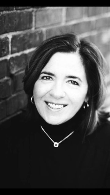 Woman smiling, wearing a black turtleneck and necklace, against a brick wall background.