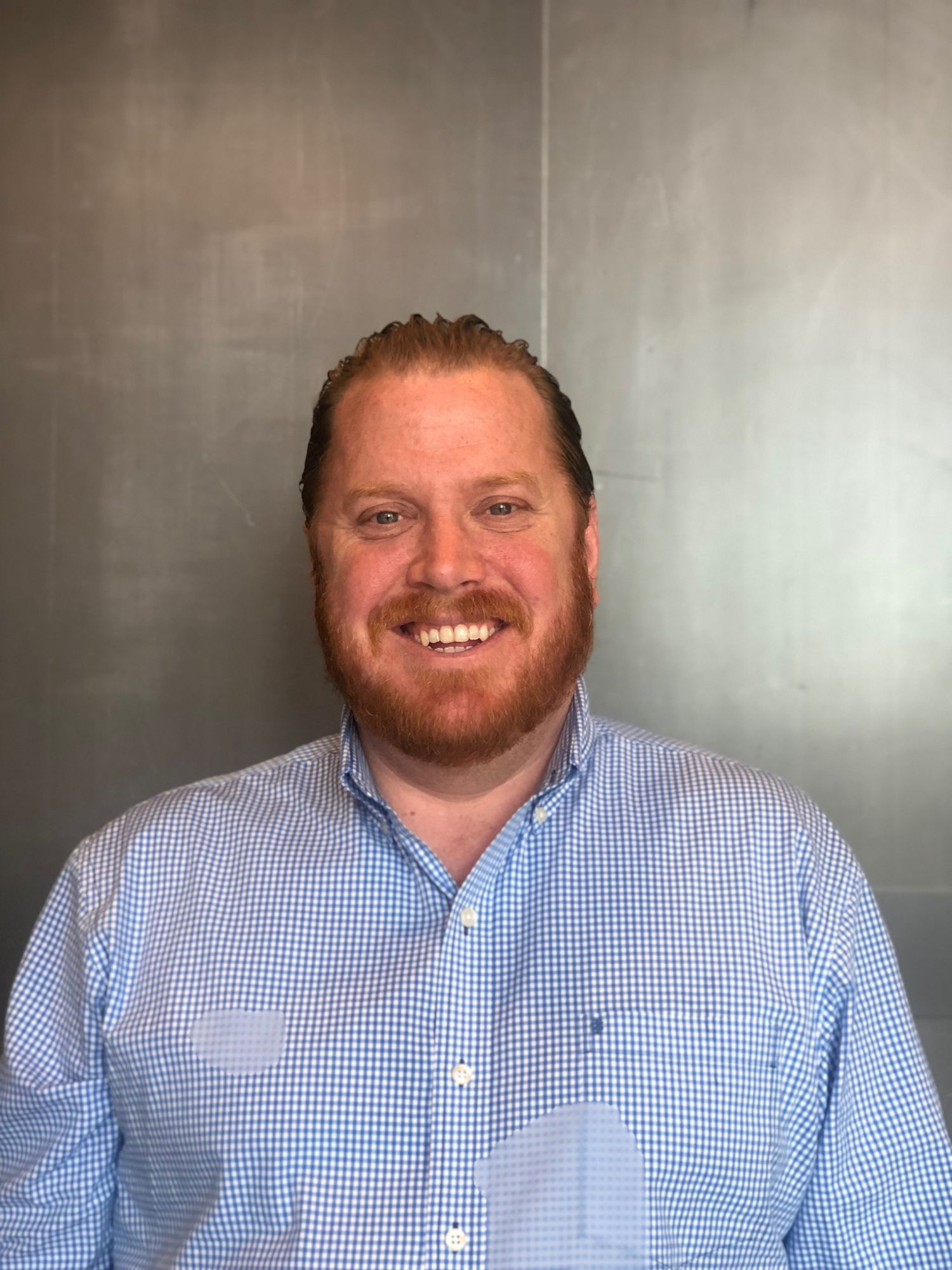 Smiling man with a beard wearing a checkered shirt against a gray background.