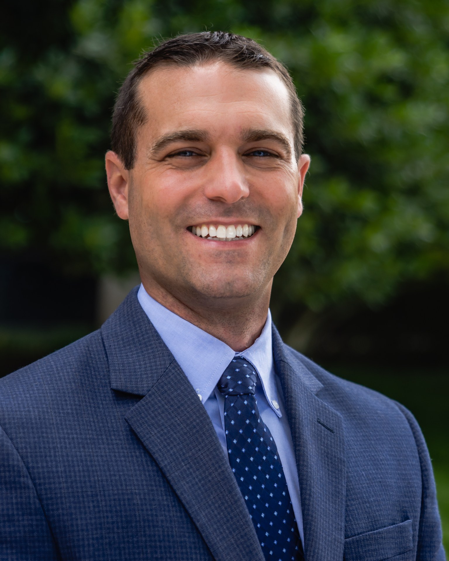 Smiling man named Peter in a suit stands outdoors with greenery in the background.