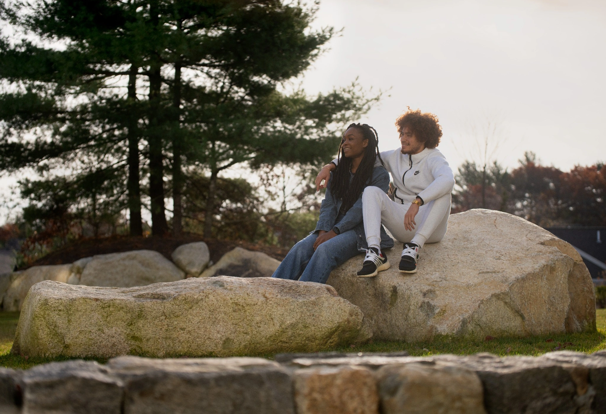 Two friends sitting on a large rock in a park, surrounded by trees.