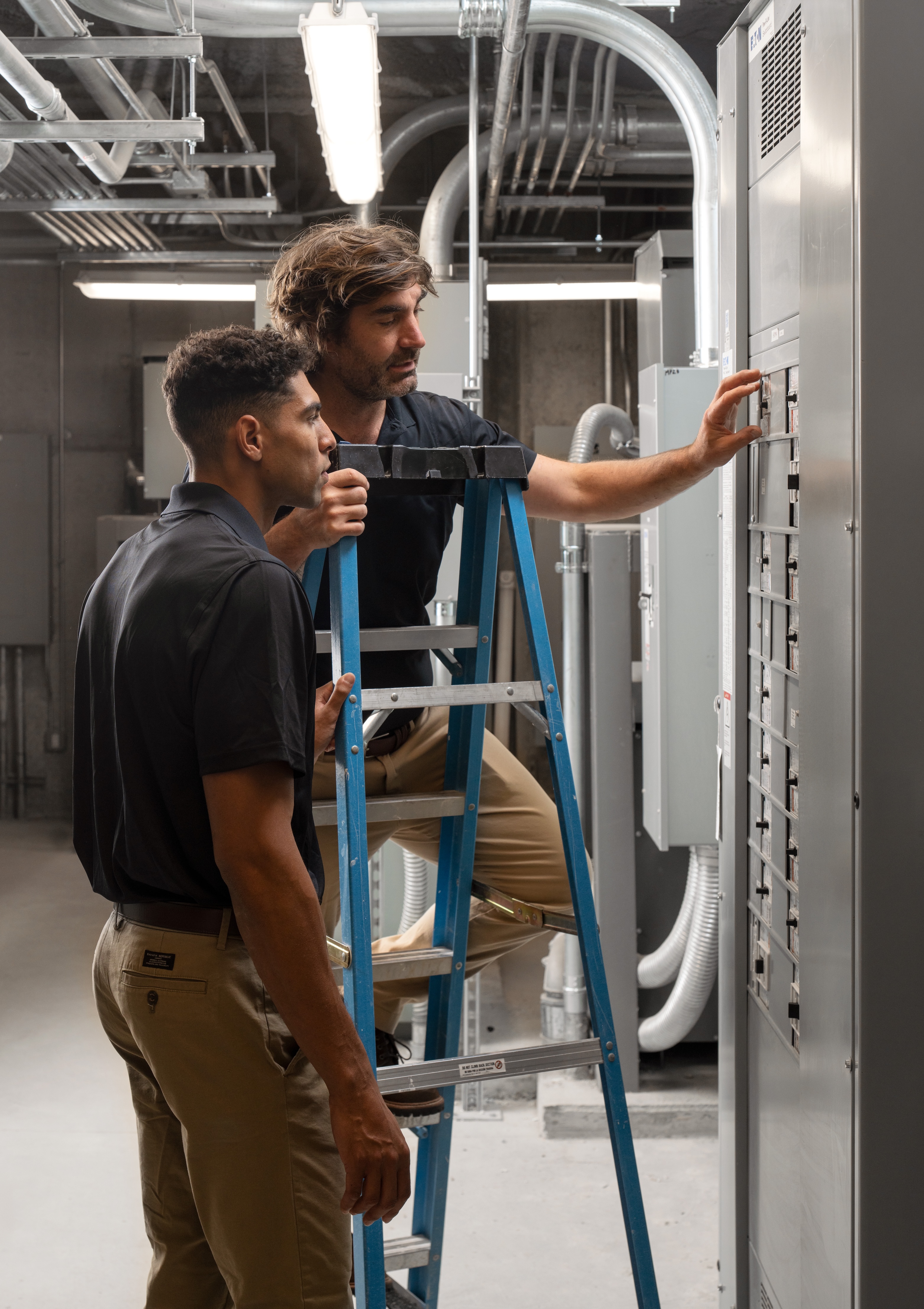 Two technicians examining a control panel, one on a ladder.