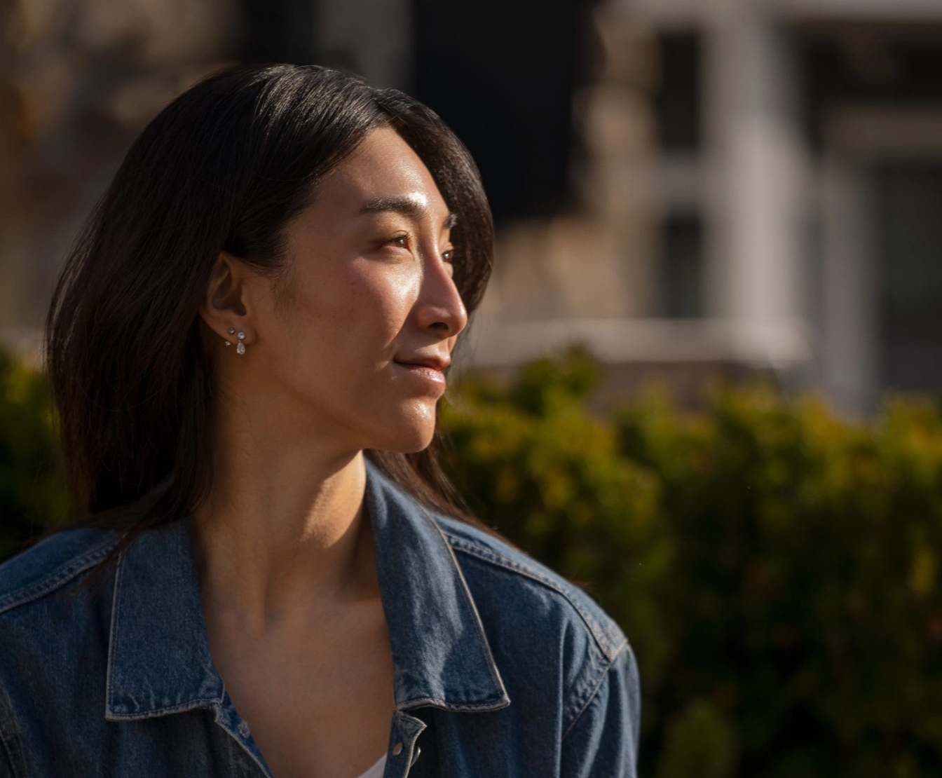 Woman with long hair wearing a denim jacket, looking to the side outdoors.
