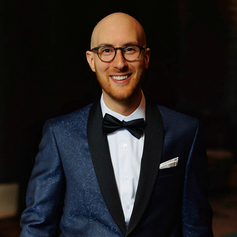 Smiling man in a tuxedo with glasses and a bow tie, standing against a dark background.