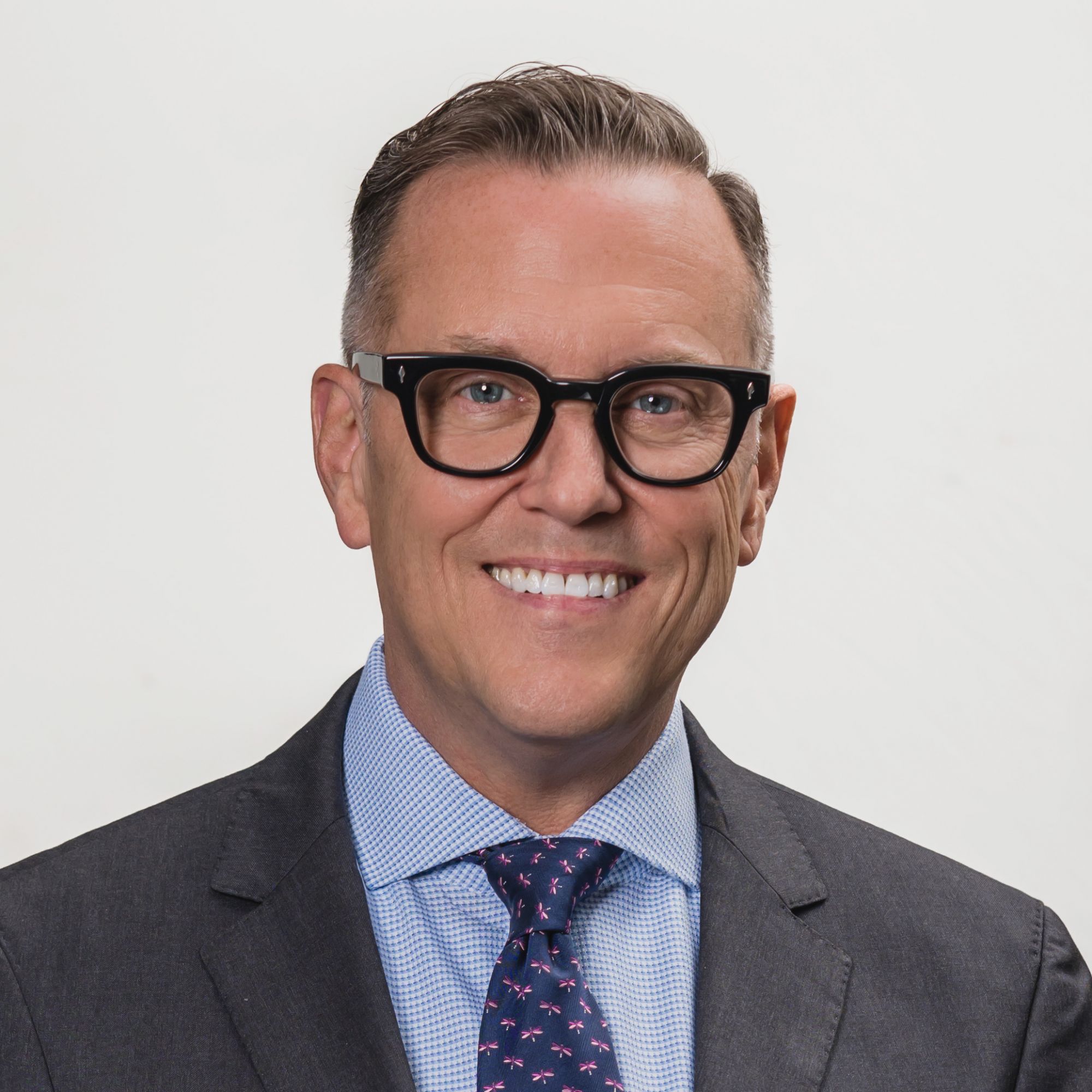 A smiling man in a suit and glasses against a plain background.
