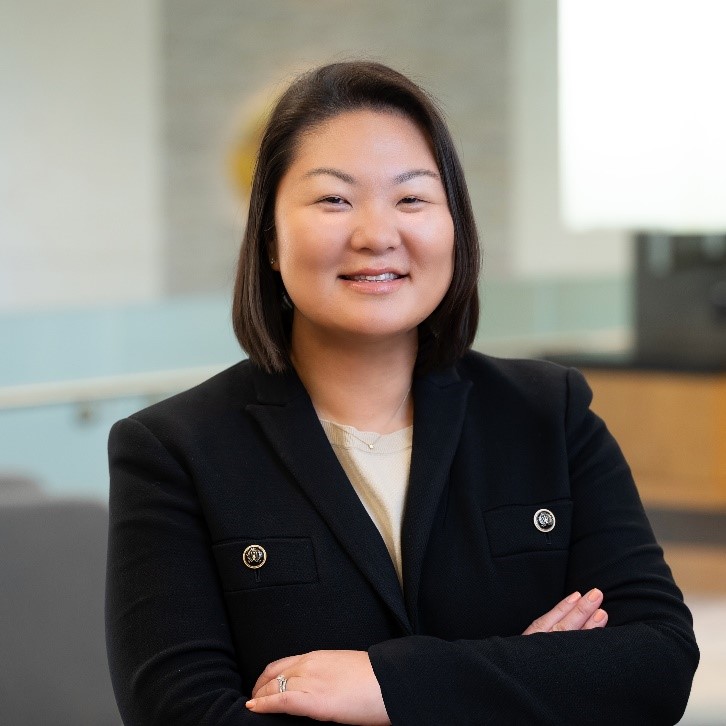 Sarah Malone smiles confidently with arms crossed, wearing a black blazer in a modern environment.