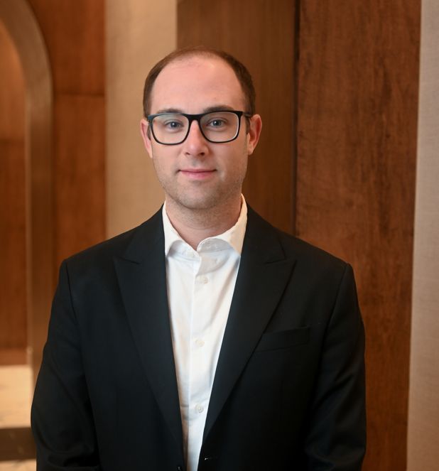 A man in glasses wearing a black suit stands in an indoor setting.