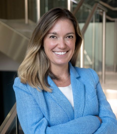 Smiling woman in a blue blazer stands confidently with arms crossed.