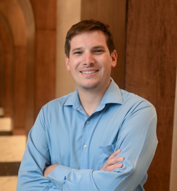 Smiling man in a blue shirt stands with arms crossed indoors.
