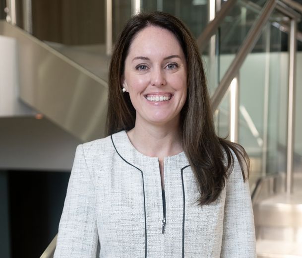 A woman in a white blazer smiles confidently in a modern office setting.