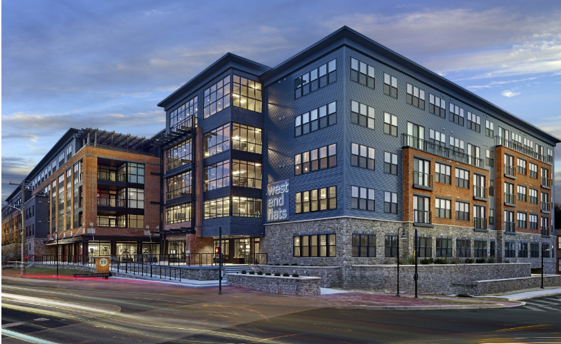 Modern mixed-use apartment building at a street corner, featuring a contemporary design with brick, stone, and blue-gray panel exteriors