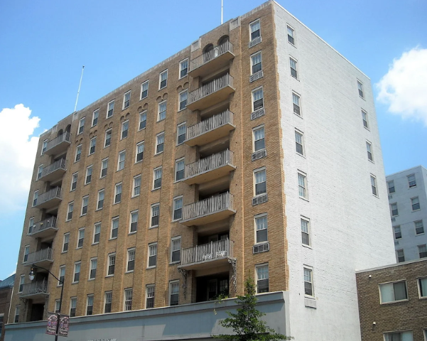 Mid-rise apartment building with a light brick exterior and a series of stacked private balconies along the front facade.