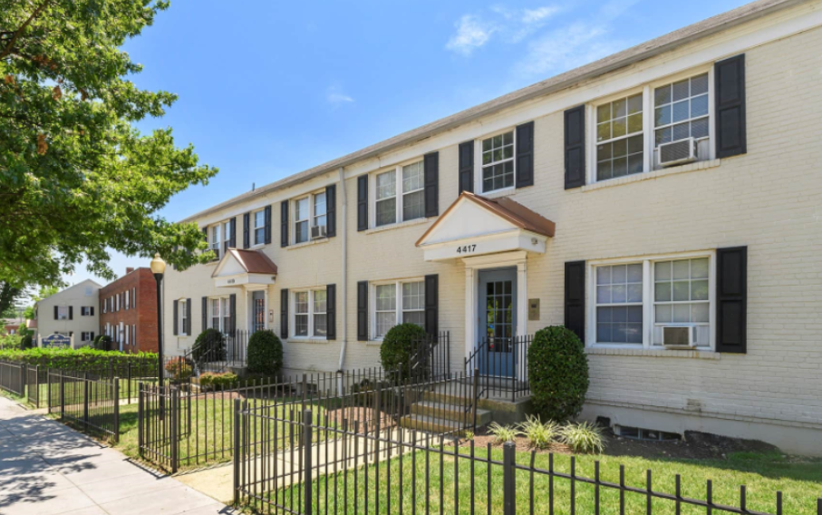 Two-story apartment building with a light brick exterior, black shutters, and covered entryways along a tree-lined sidewalk