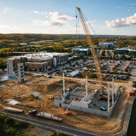 Aerial view of an active construction site with a tower crane, concrete walls and columns, and surrounding buildings and autumn foliage.