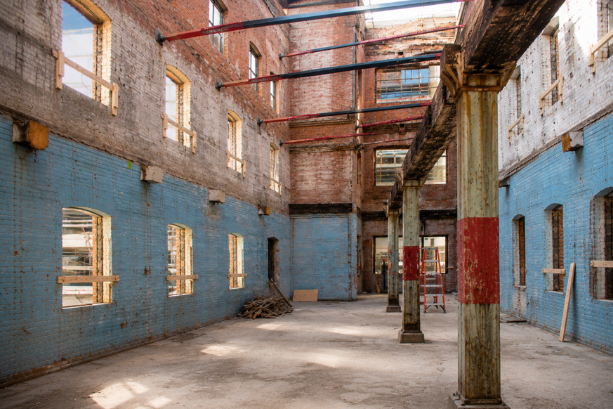 Interior of a historic brick building under renovation, with exposed steel beams, columns, open windows, and blue-painted walls.