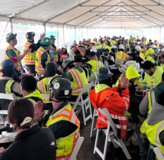 Construction workers gathered for a meal and presentation under a large event tent.