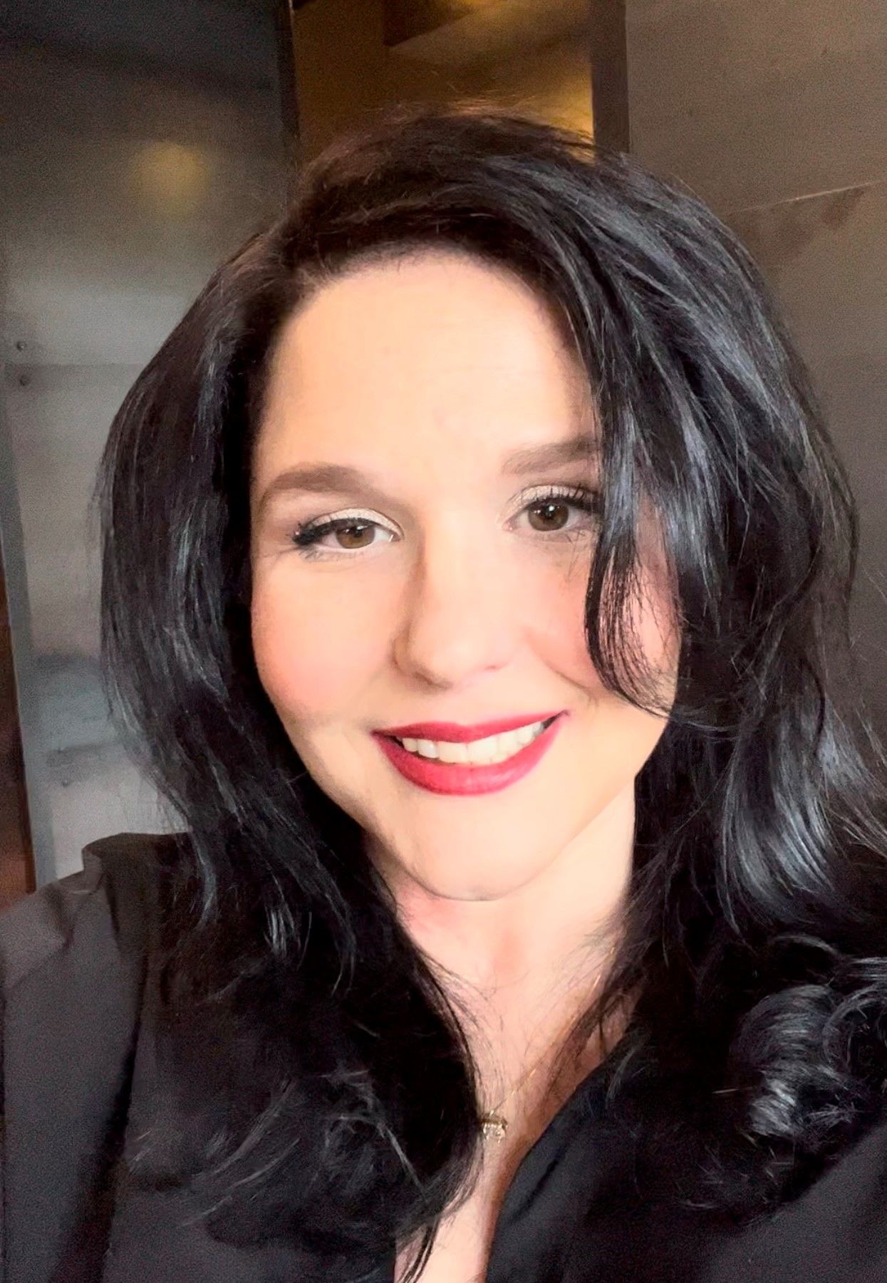 A woman with long black hair smiles, wearing a black top and subtle makeup.