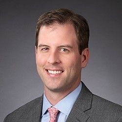 Male professional in a suit with a light blue shirt and pink tie, smiling against a gray background.