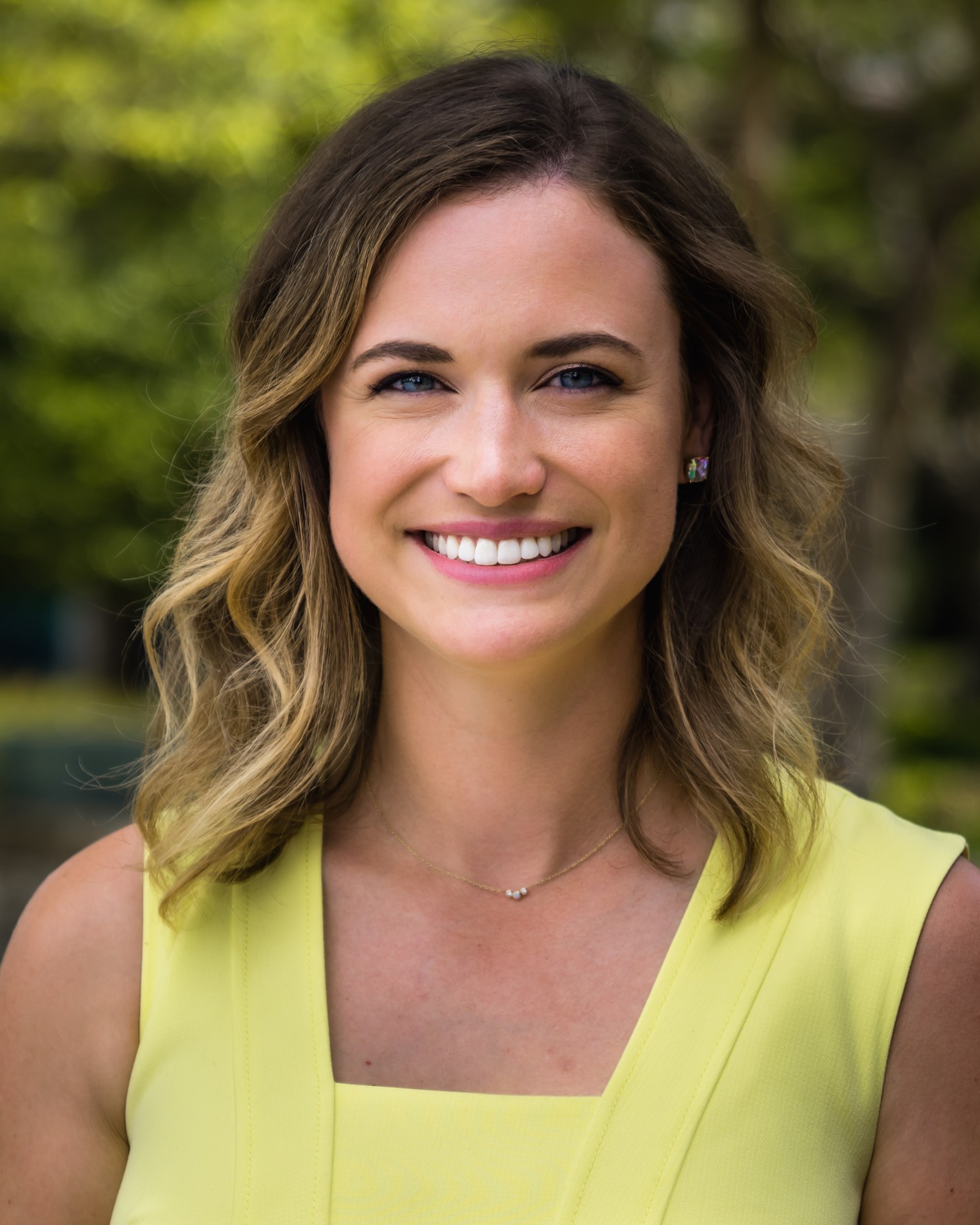 Tess Guinn smiling in a yellow dress with greenery in the background.
