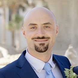 Man in a blue suit with a light tie, smiling outdoors with greenery in the background.