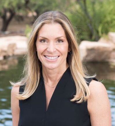 A woman with long blonde hair smiles, standing by a serene water backdrop.