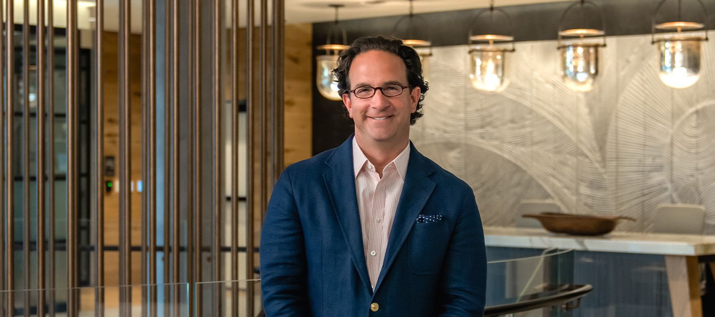 A smiling man in a blazer stands in a modern, well-lit interior space.