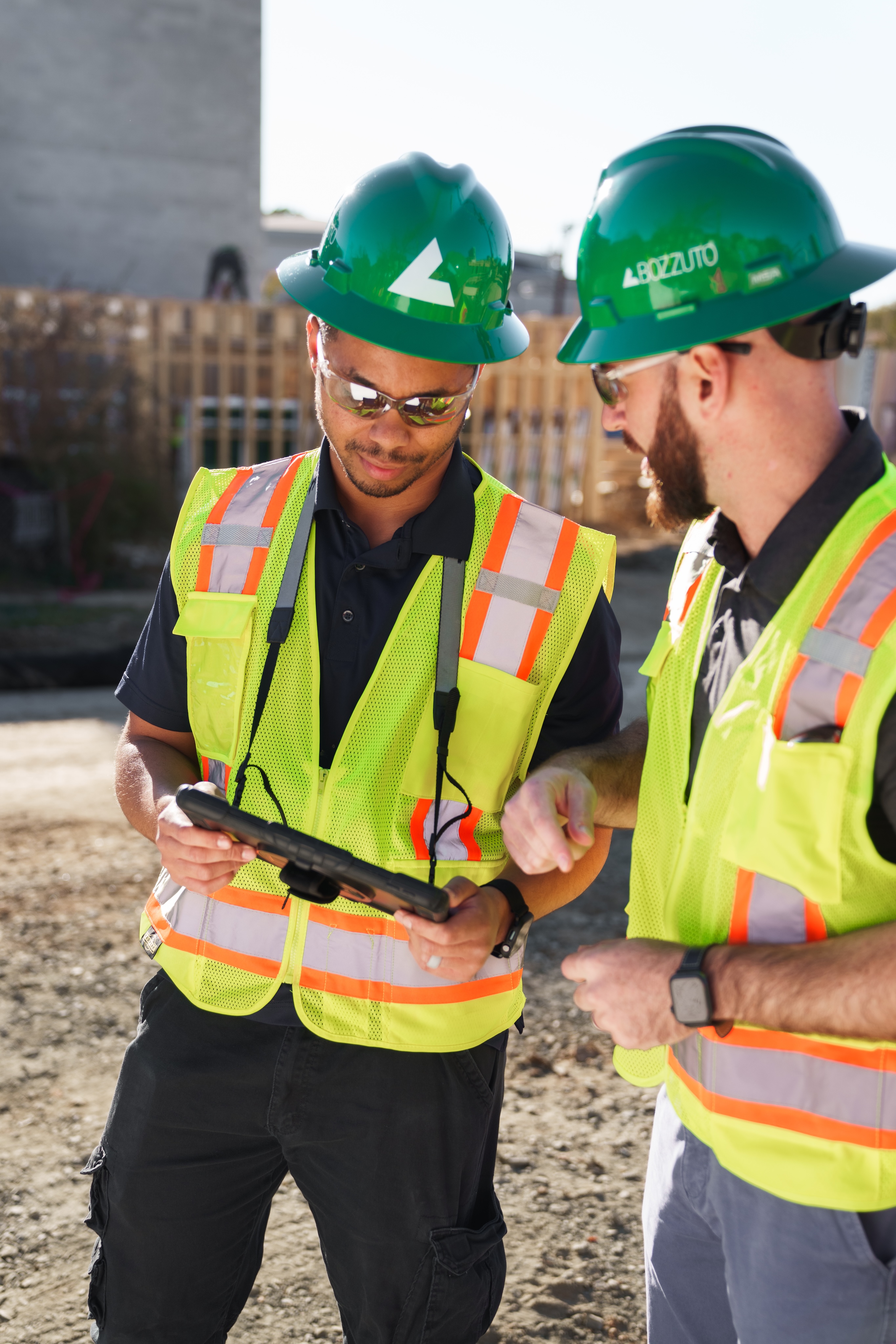 Two construction workers in safety vests and helmets inspecting a tablet.