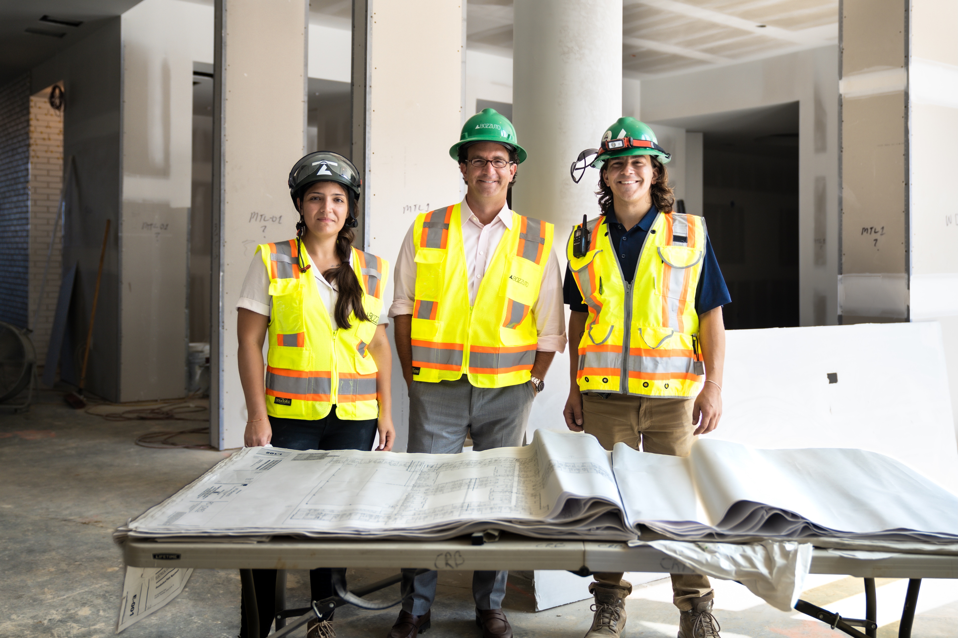 Three individuals in hard hats and safety vests stand by a table of blueprints.