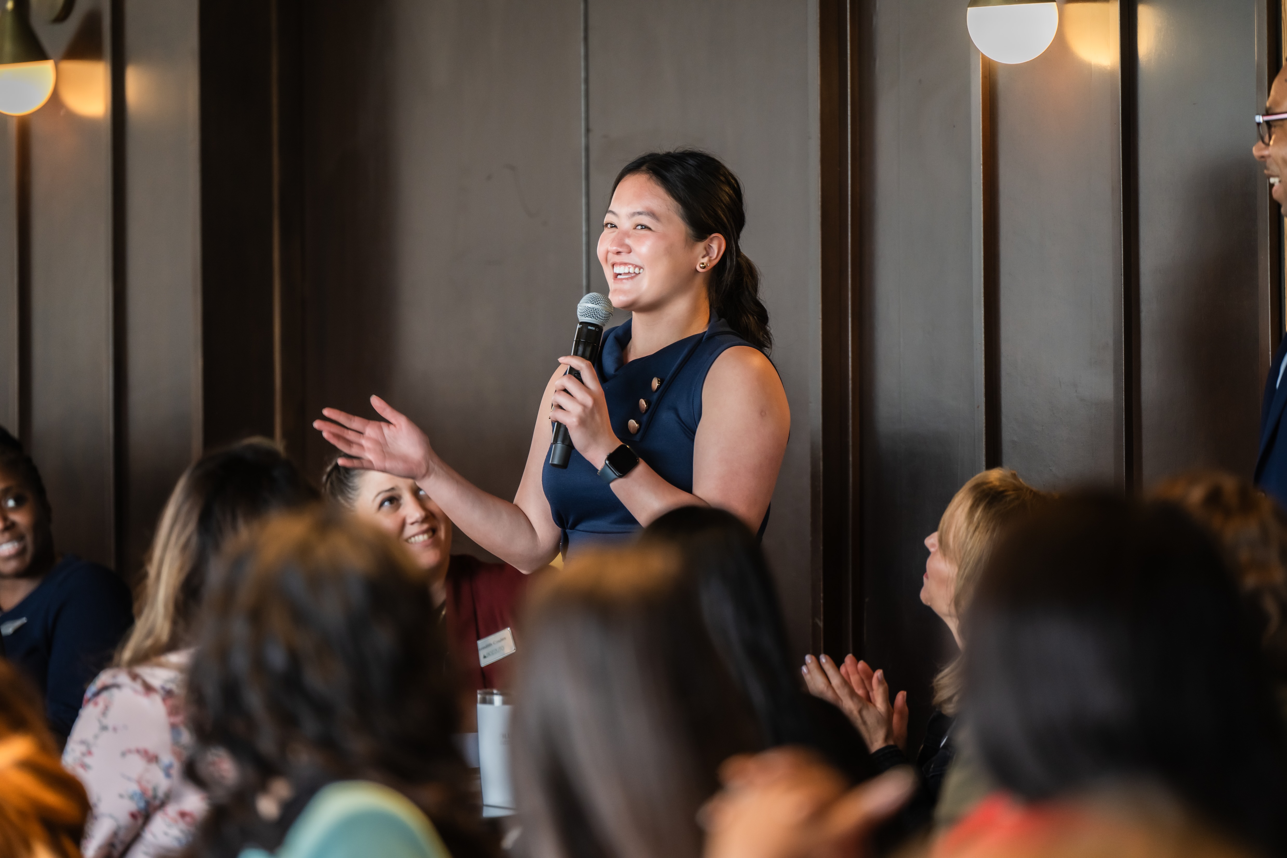 Woman speaking into a microphone at a gathering, audience engaged.