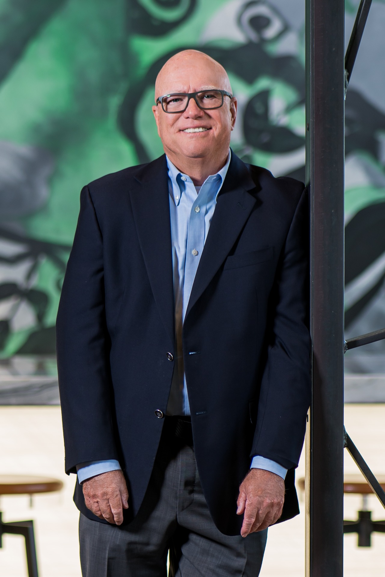 Richard L. Mostyn stands in front of a colorful mural, wearing a blazer and smiling.