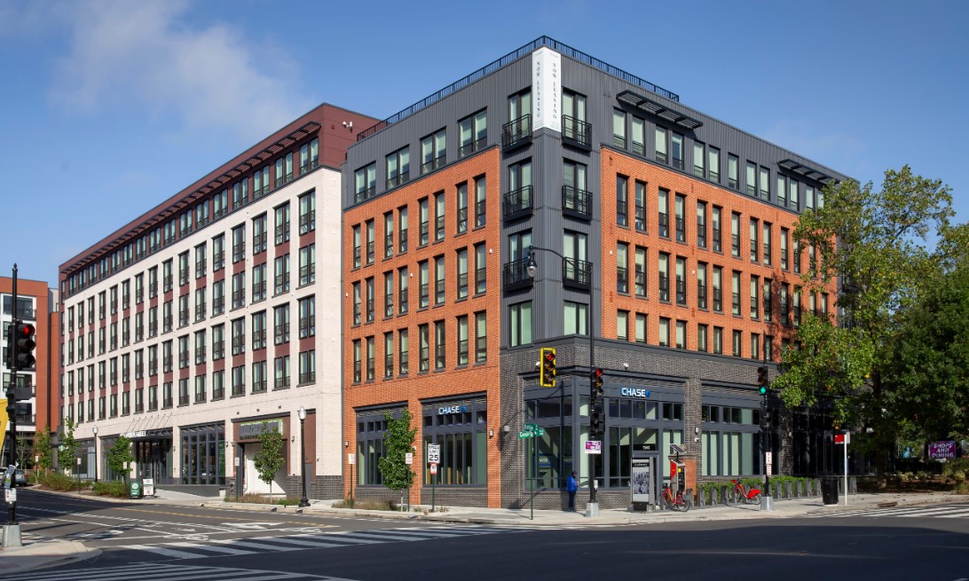 Modern mixed-use apartment building at a prominent city street corner, featuring a red brick, white, and dark metal exterior with large windows and balconies.