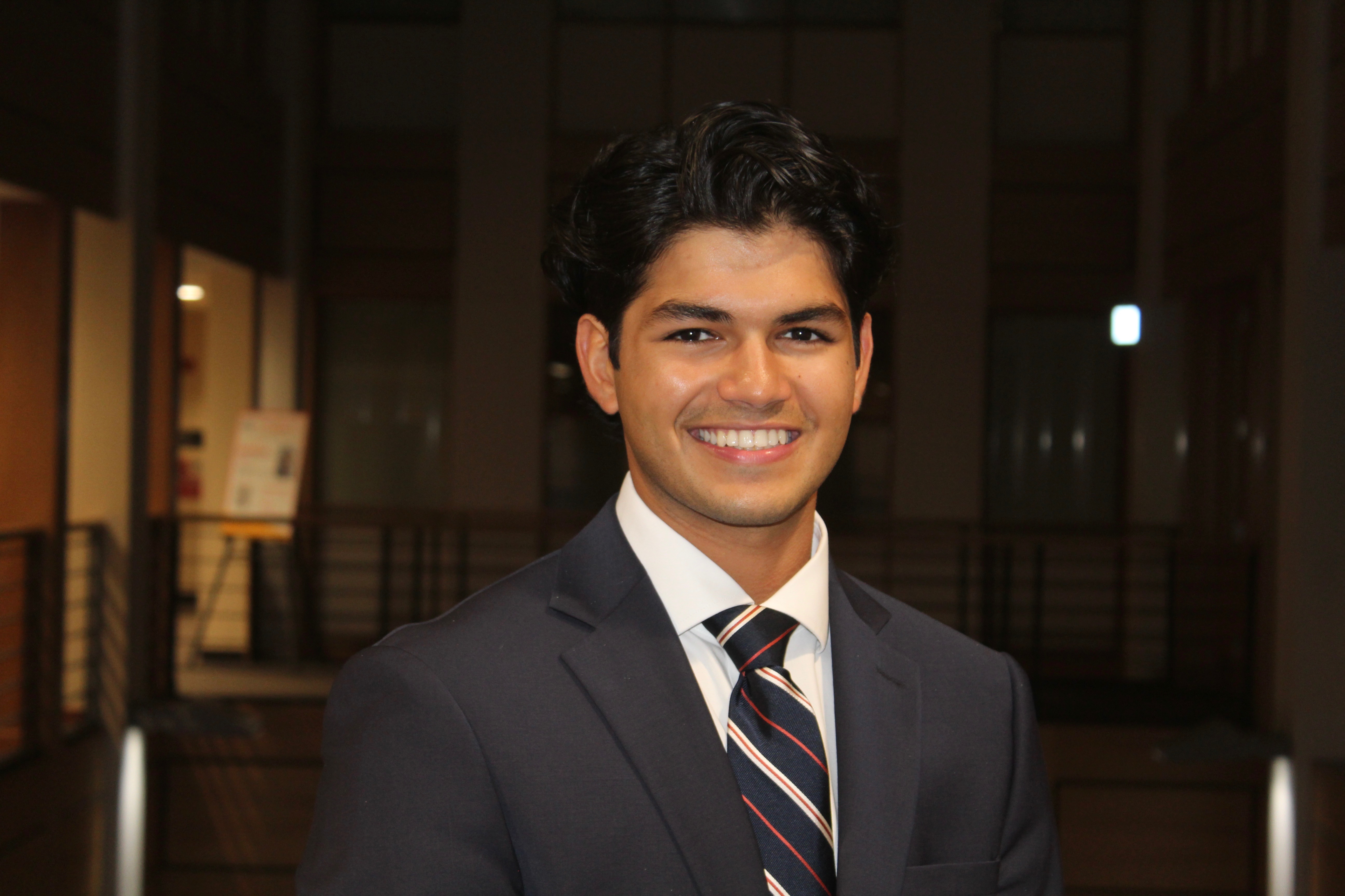 A young man smiling in a suit with a tie, standing indoors.