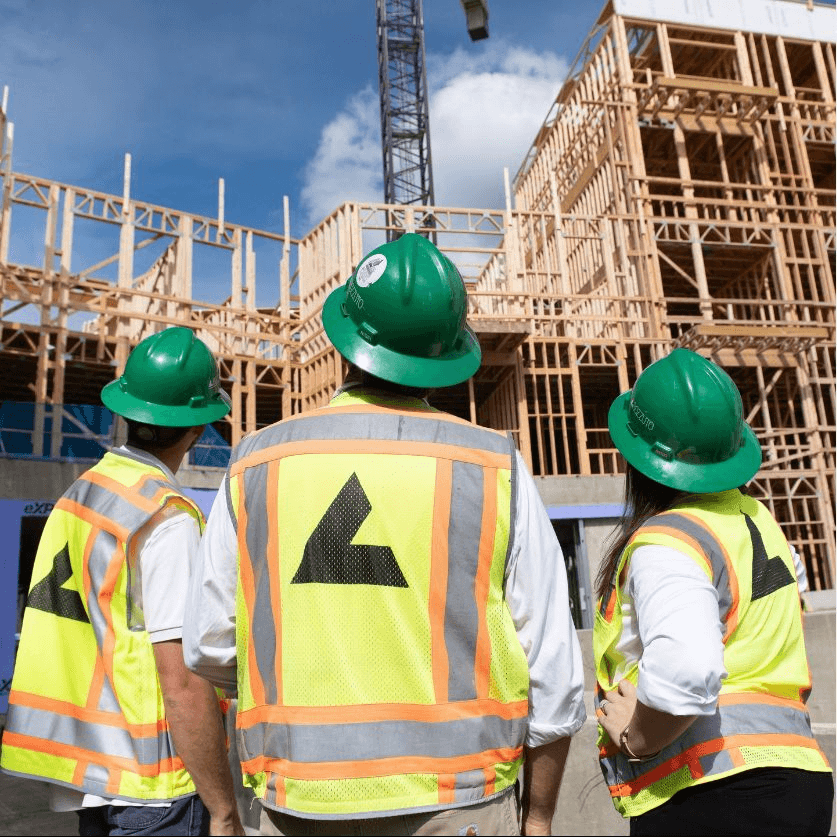 Three construction workers in hard hats observe a building under construction.