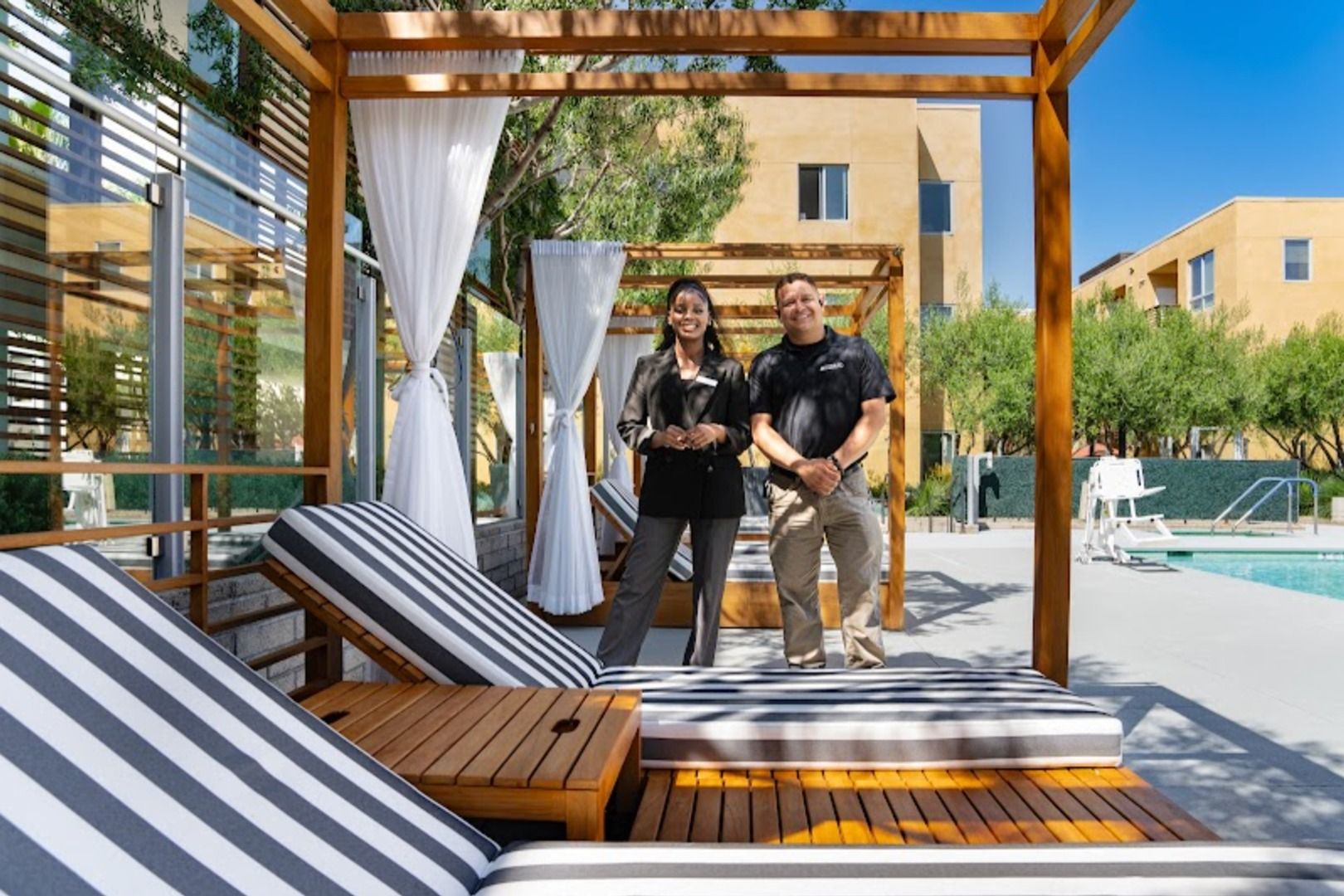 Two people stand by lounge chairs near a pool surrounded by trees and buildings.