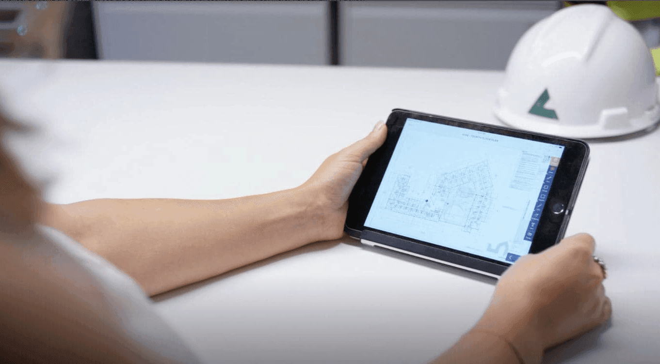 Person holding a tablet displaying a technical plan, with a hard hat in the background.