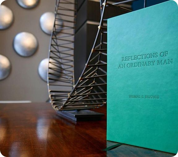 A teal book titled "Reflections of an Ordinary Man" beside a modern sculpture on a wooden table.