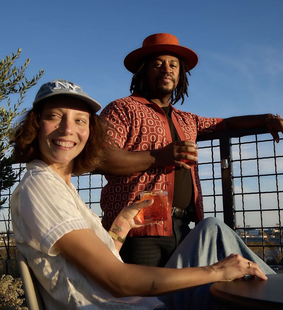 A woman and a man smile while holding drinks against a sunset backdrop.