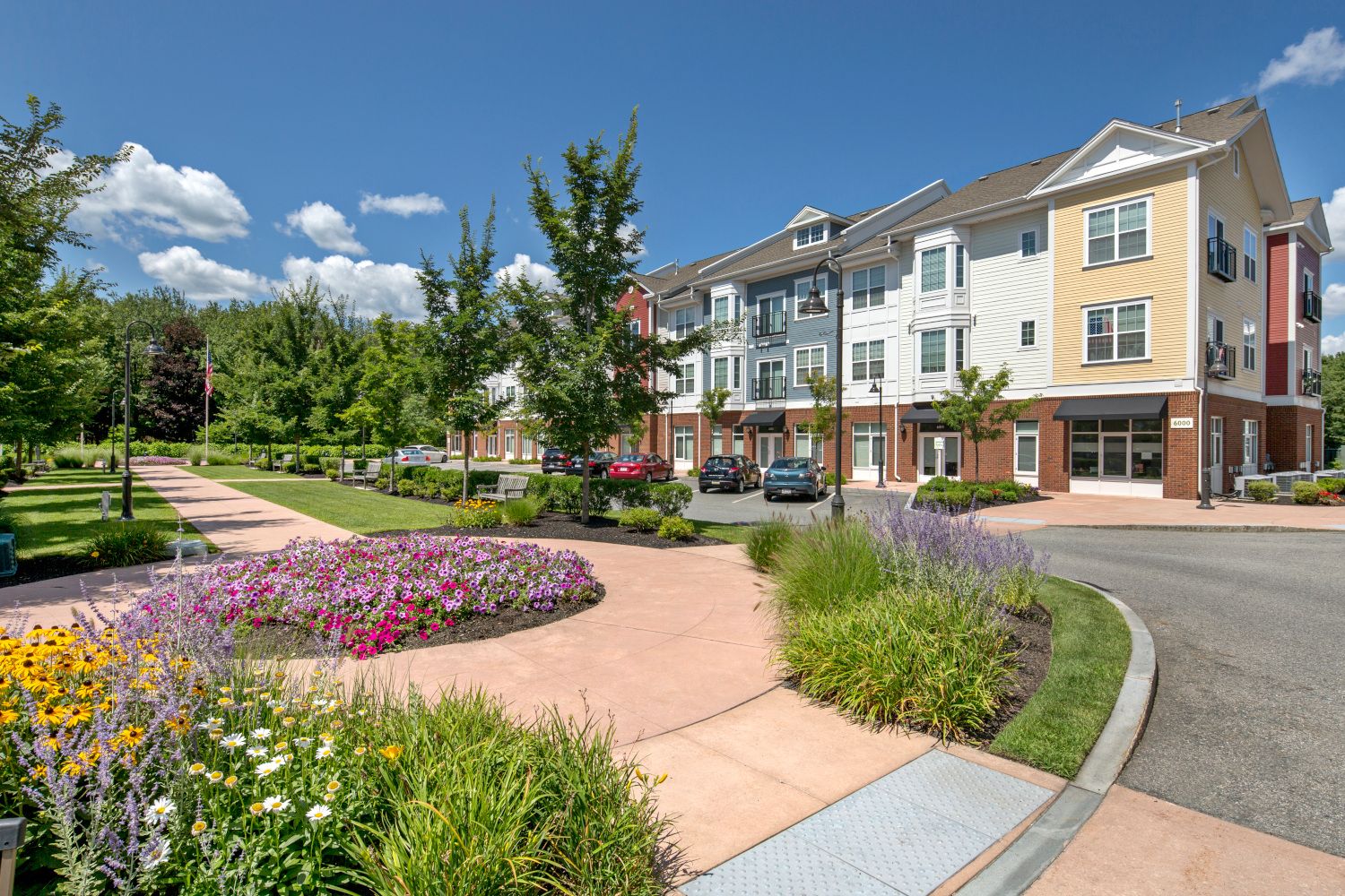 Colorful flower beds line a pathway beside modern townhouses under a blue sky.