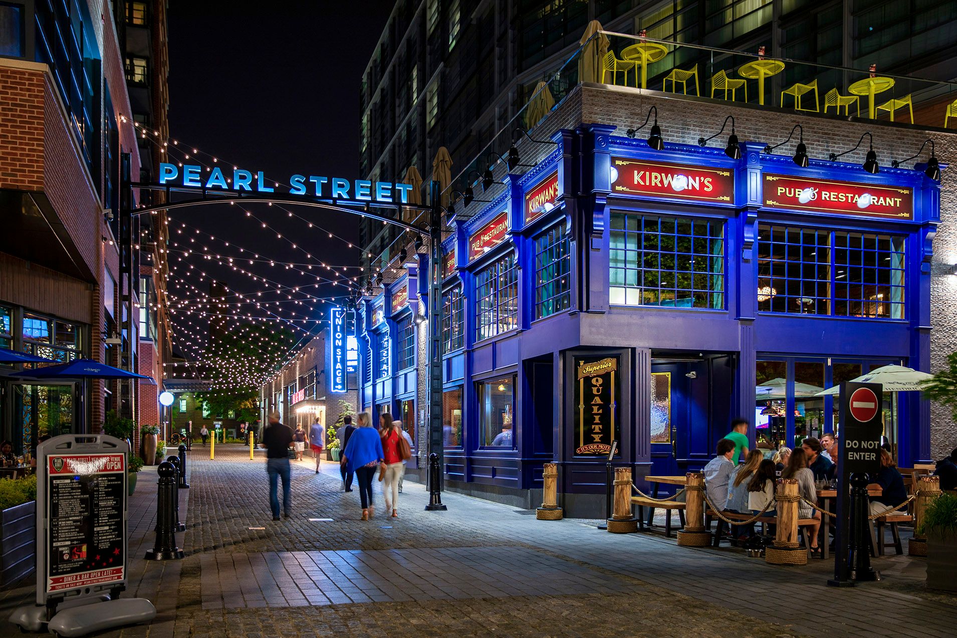 Night scene of a lively street with illuminated buildings and people dining outside.