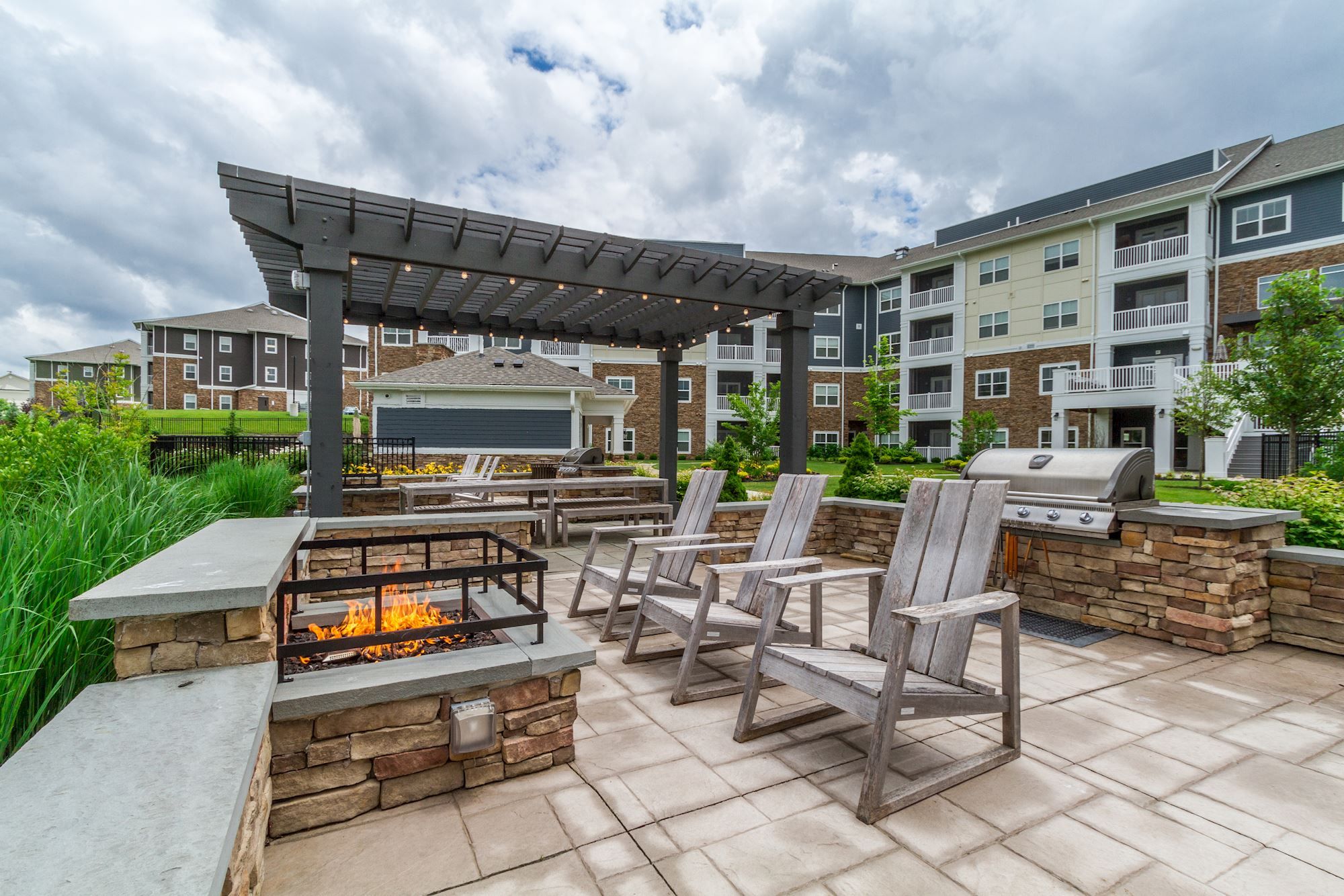 Outdoor seating area with a fire pit and grill, surrounded by greenery and buildings.