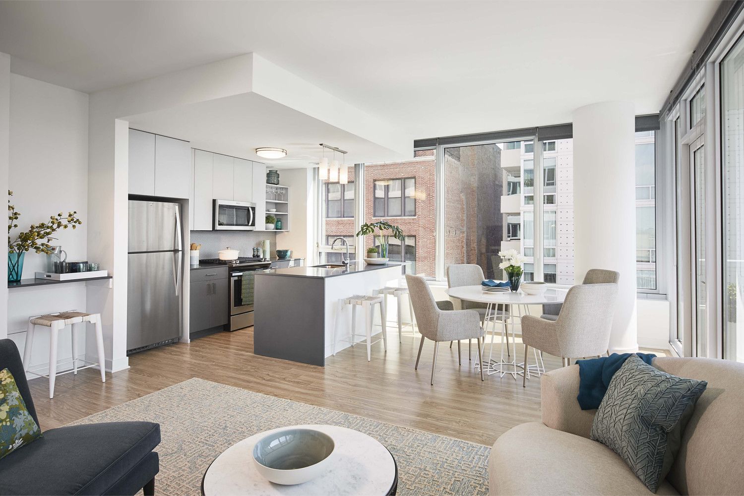 Modern kitchen and living area with light tones and large windows, showcasing city views.