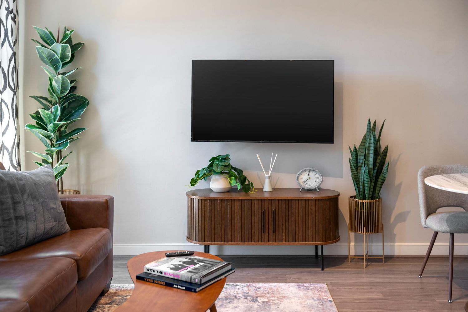 Apartment TV wall at 77 H with mounted TV, wood media console, plants, sofa edge, and coffee table