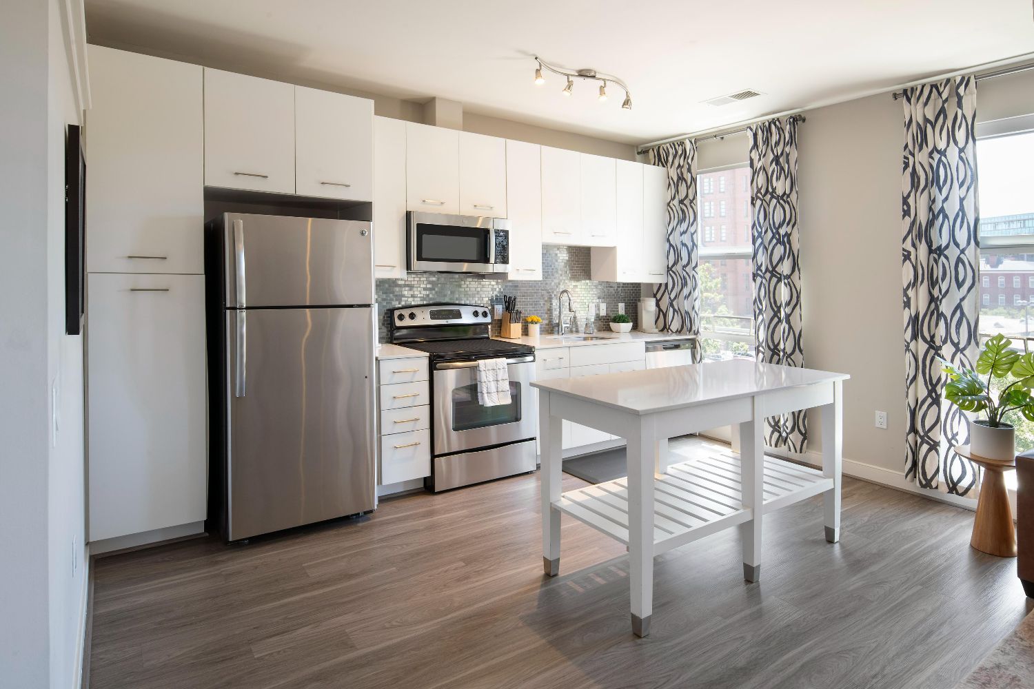 Apartment kitchen at 77 H with white island, stainless steel refrigerator, range, microwave, patterned curtains, and bright windows