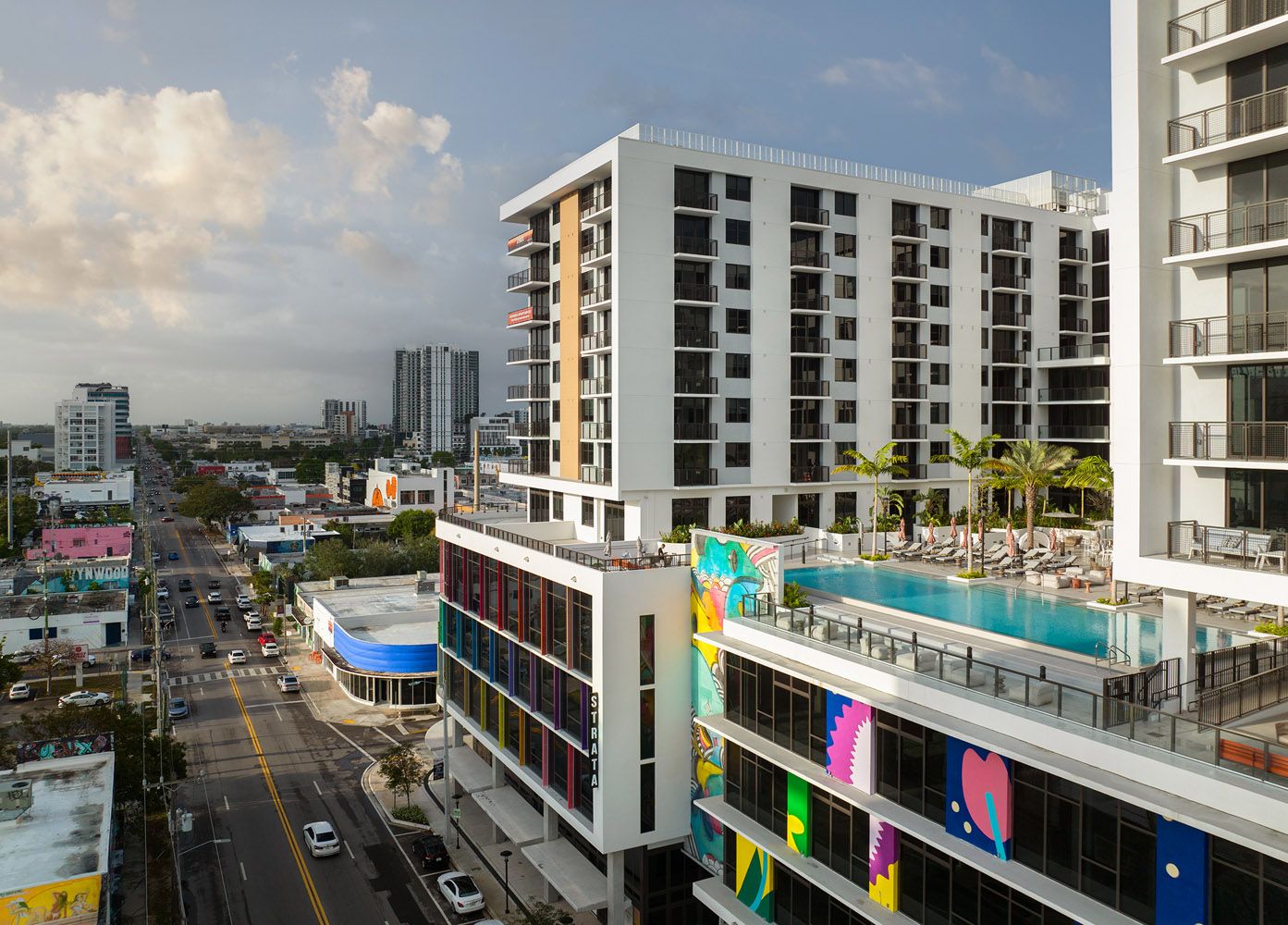 Modern multi-story building with a rooftop pool and colorful murals overlooking a city street.